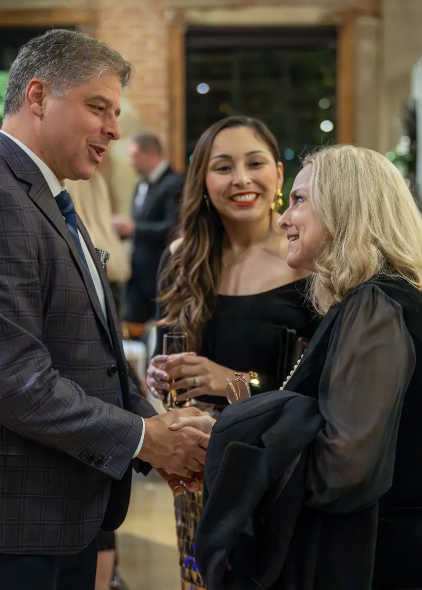 The image shows three people engaged in a warm, conversational moment at what appears to be a formal or semi‑formal event.